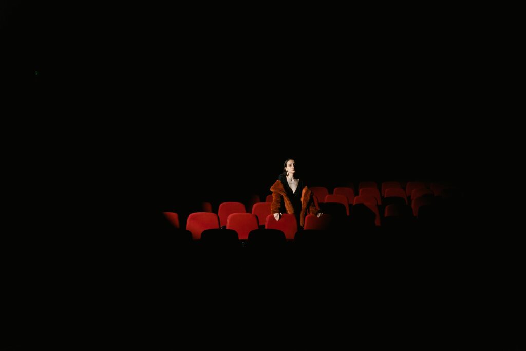 A woman in a fur coat sits alone in a dimly lit theater, surrounded by empty red seats.